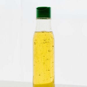 Yellow body oil with bubbles in transparent plastic bottle with green cap on table against white background