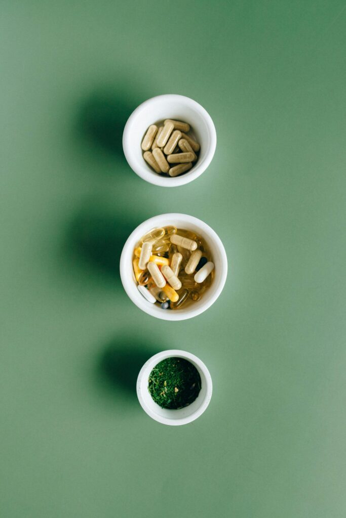 Top view of bowls with capsules, herbs, and supplements on green surface for healthcare concept.